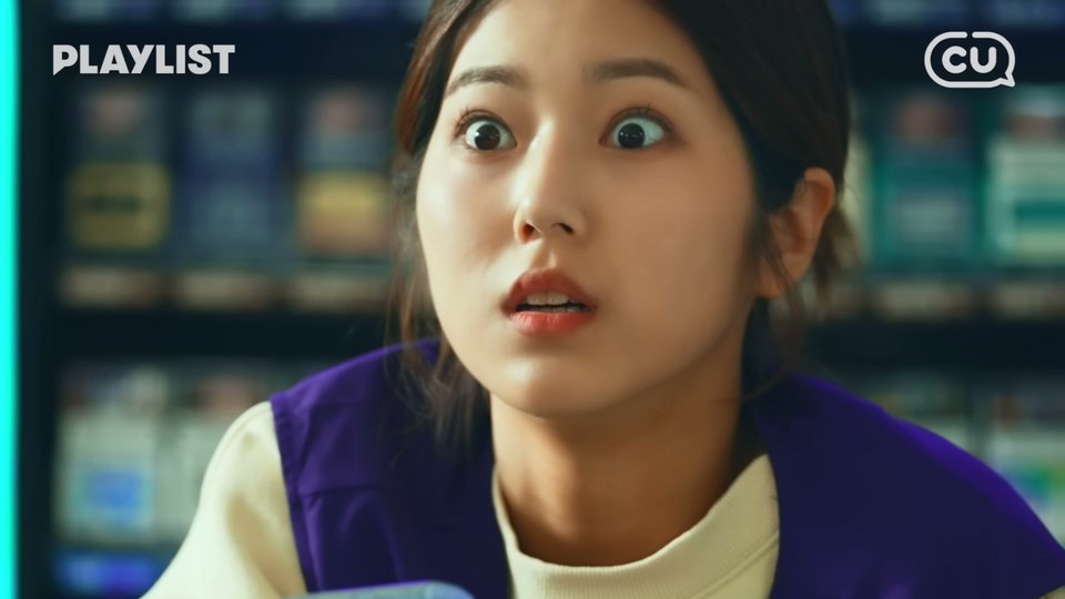 보라색 조끼를 입은 알바생이 무늬 케이스 휴대폰을 쥐고 눈을 크게 뜬 채 앞을 보고, 뒤로 담배 진열대가 흐리게 보이는 장면