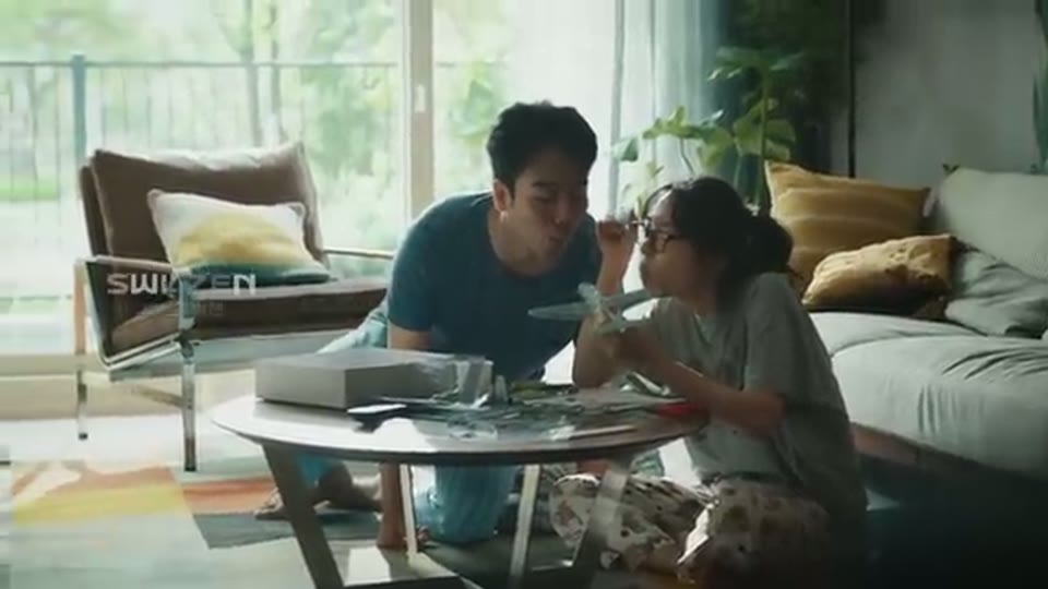 햇빛이 드는 거실에서 두 사람이 낮은 테이블에 마주 앉아 함께 음식을 먹고 왼쪽 의자 뒤에 브랜드 로고가 보이는 장면