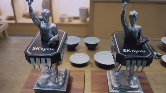 나무 탁자 위 흰 도자기 그릇들 사이에 칩 모양 받침에 SK hynix 로고가 새겨진 사람 모양 트로피 두 개가 놓인 장면